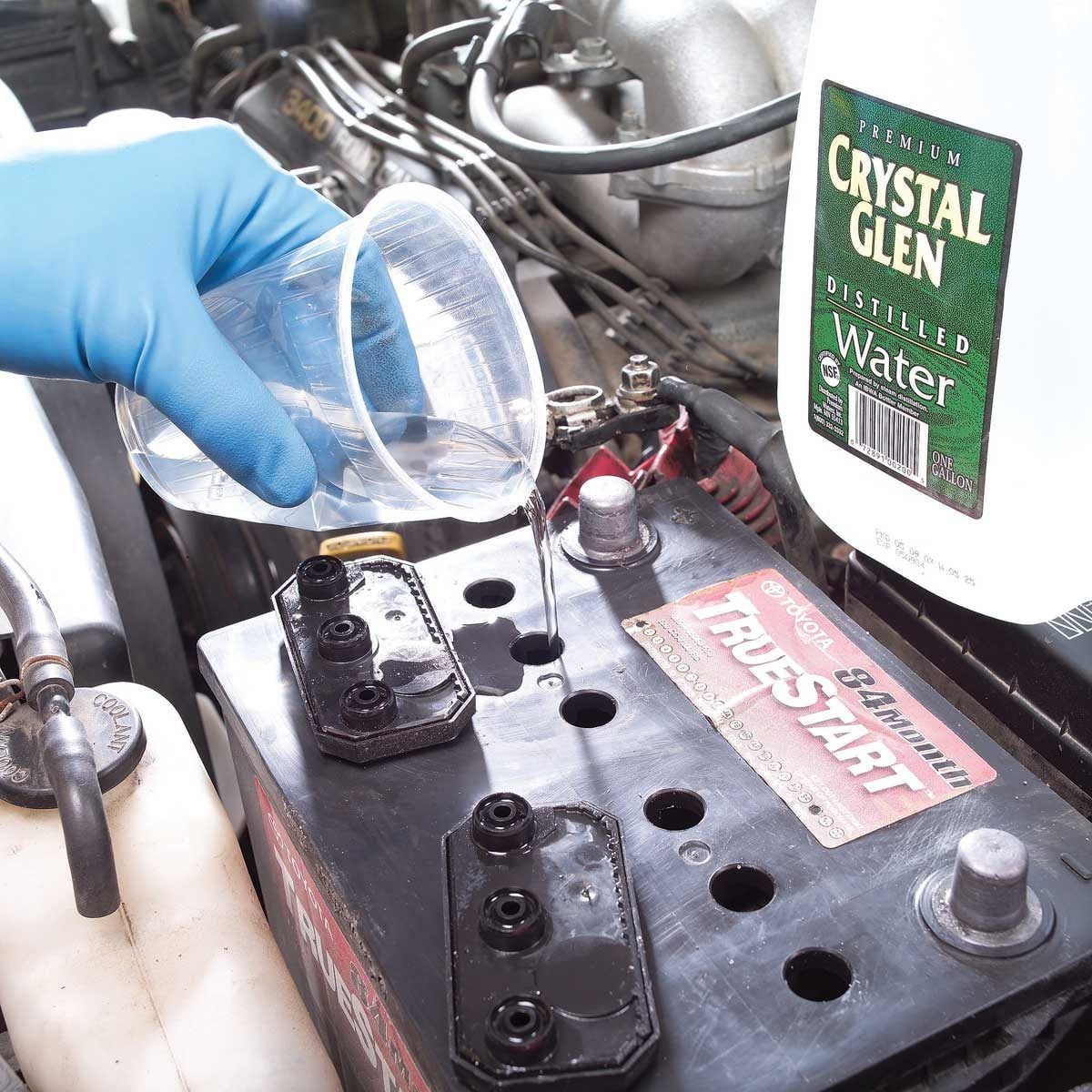 A gloved hand pours distilled water from a cup into a car battery, surrounded by engine components and a gallon of Crystal Glen water.