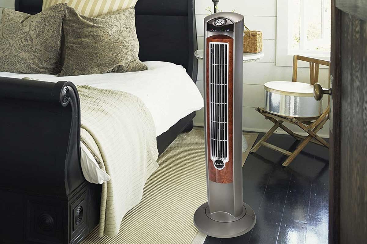 A tall, sleek fan stands in a bedroom, providing airflow beside a bed with decorative pillows and a chair in a well-lit, cozy environment.