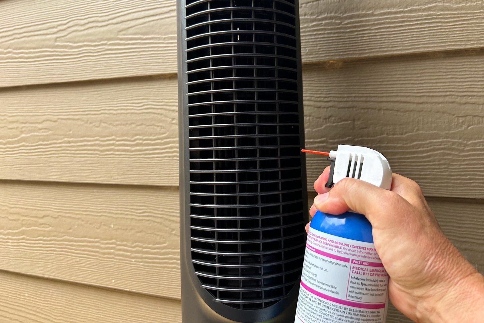 A hand holds a compressed air canister aimed at a black air vent, cleaning it while positioned against a textured beige wall.
