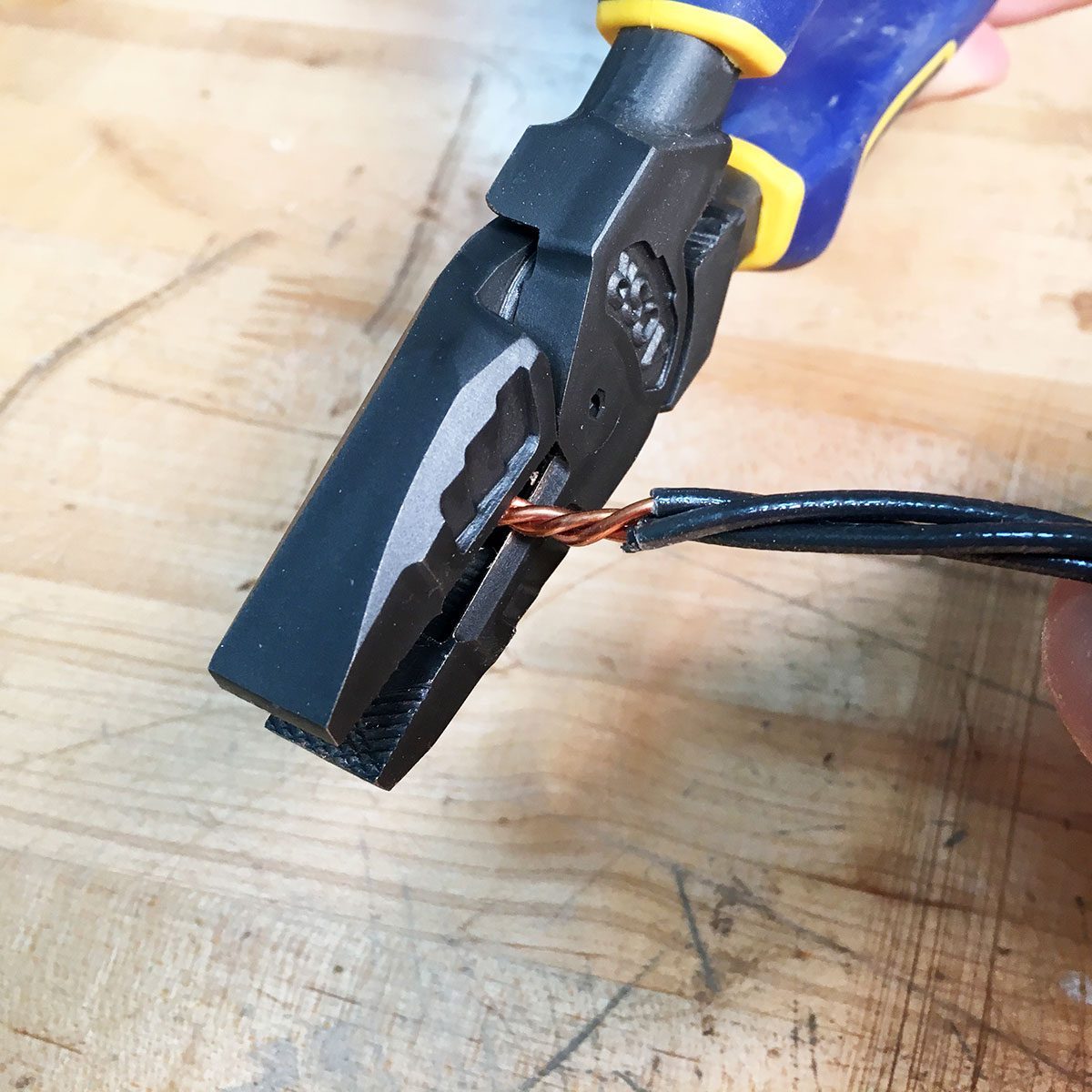 Clipping wires with linesmans pliers. A pair of wire cutters grips a twisted copper wire, positioned on a wooden surface, indicating preparation for cutting or stripping the wire.
