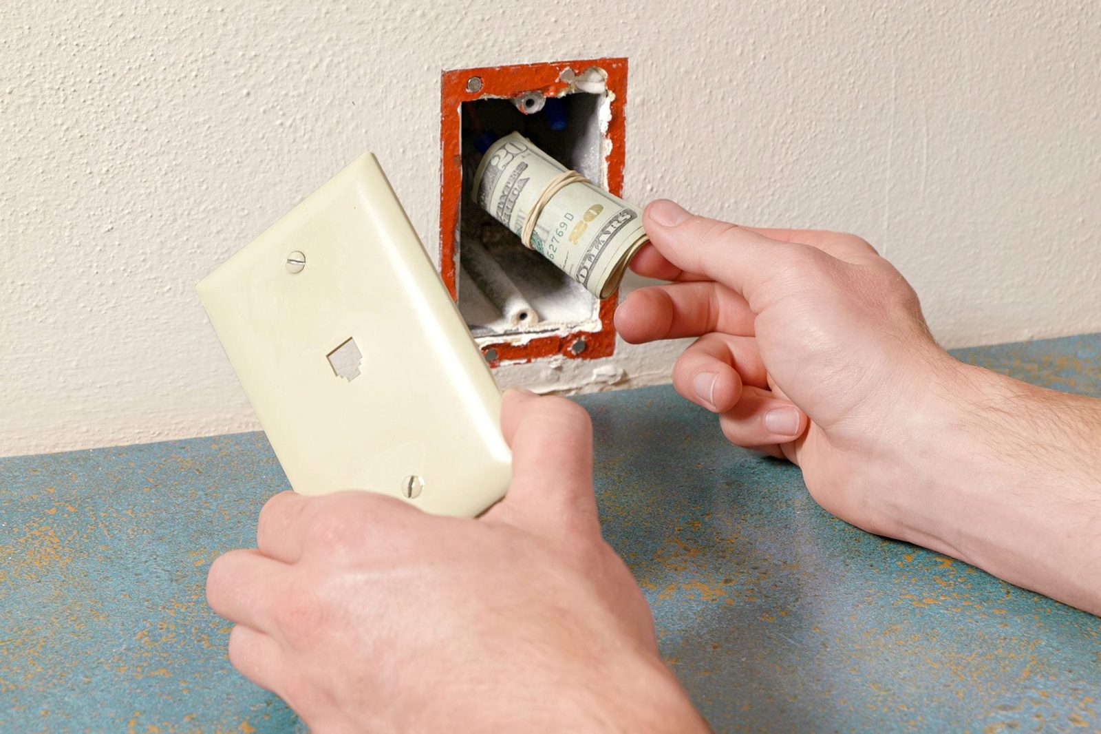 a close-up of a person's hands removing a light switch cover, Behind the switch, there’s a rolled-up banknote hidden inside the electrical b