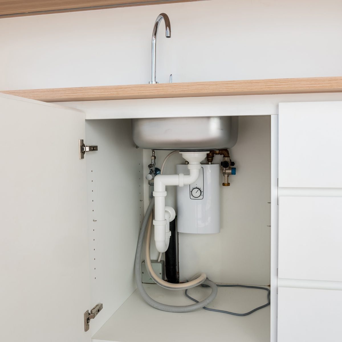 A white sink cabinet contains plumbing components; pipes connect to a filtration unit, situated below a stainless steel sink in a modern kit