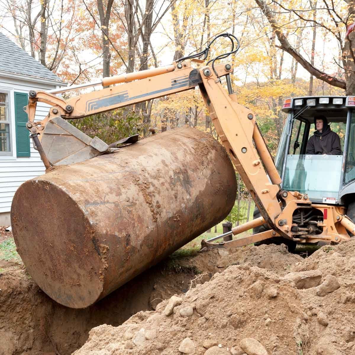A backhoe excavator lifts a large, rusty tank from a dug-out area beside a house, with autumn trees in the background.