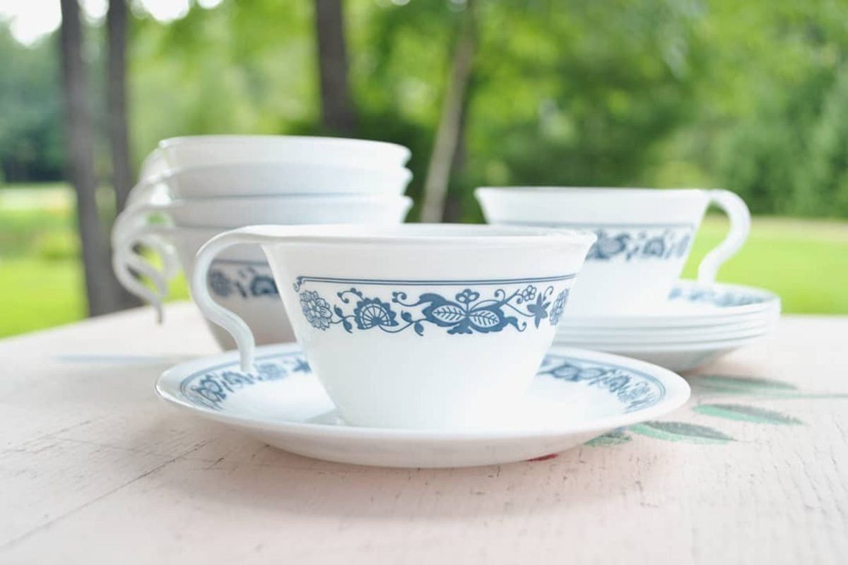 A white tea cup and saucer, adorned with blue floral designs, sit on a wooden table, surrounded by stacked cups, against a blurred green bac