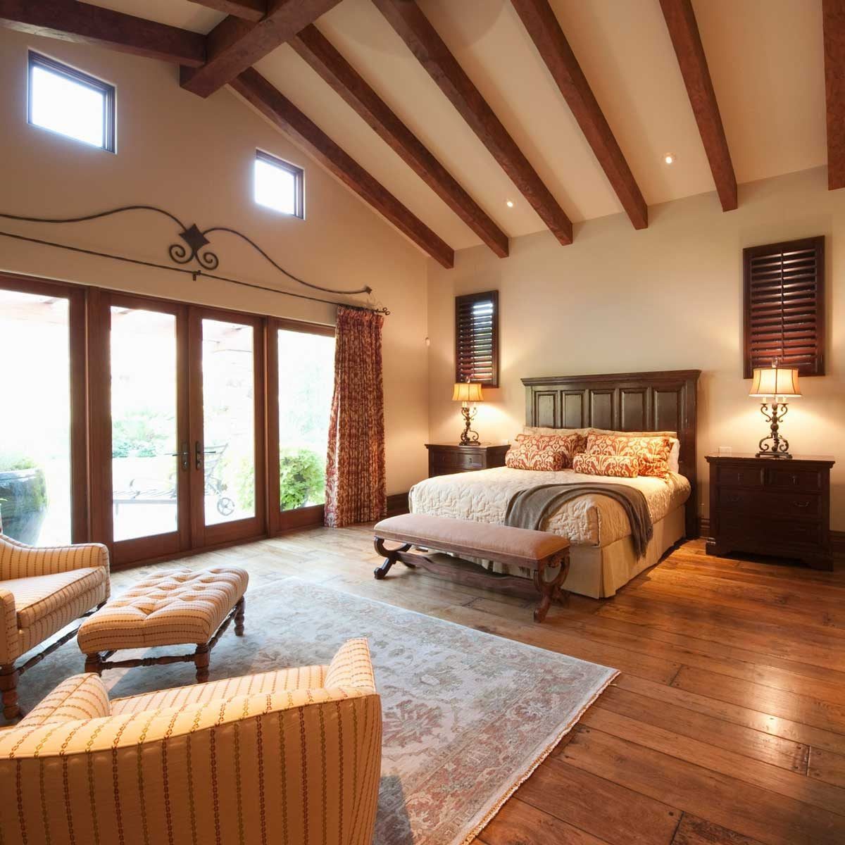 A furnished bedroom features a large bed with decorative pillows, a small bench at its foot, and cozy seating by a glass door leading outsid