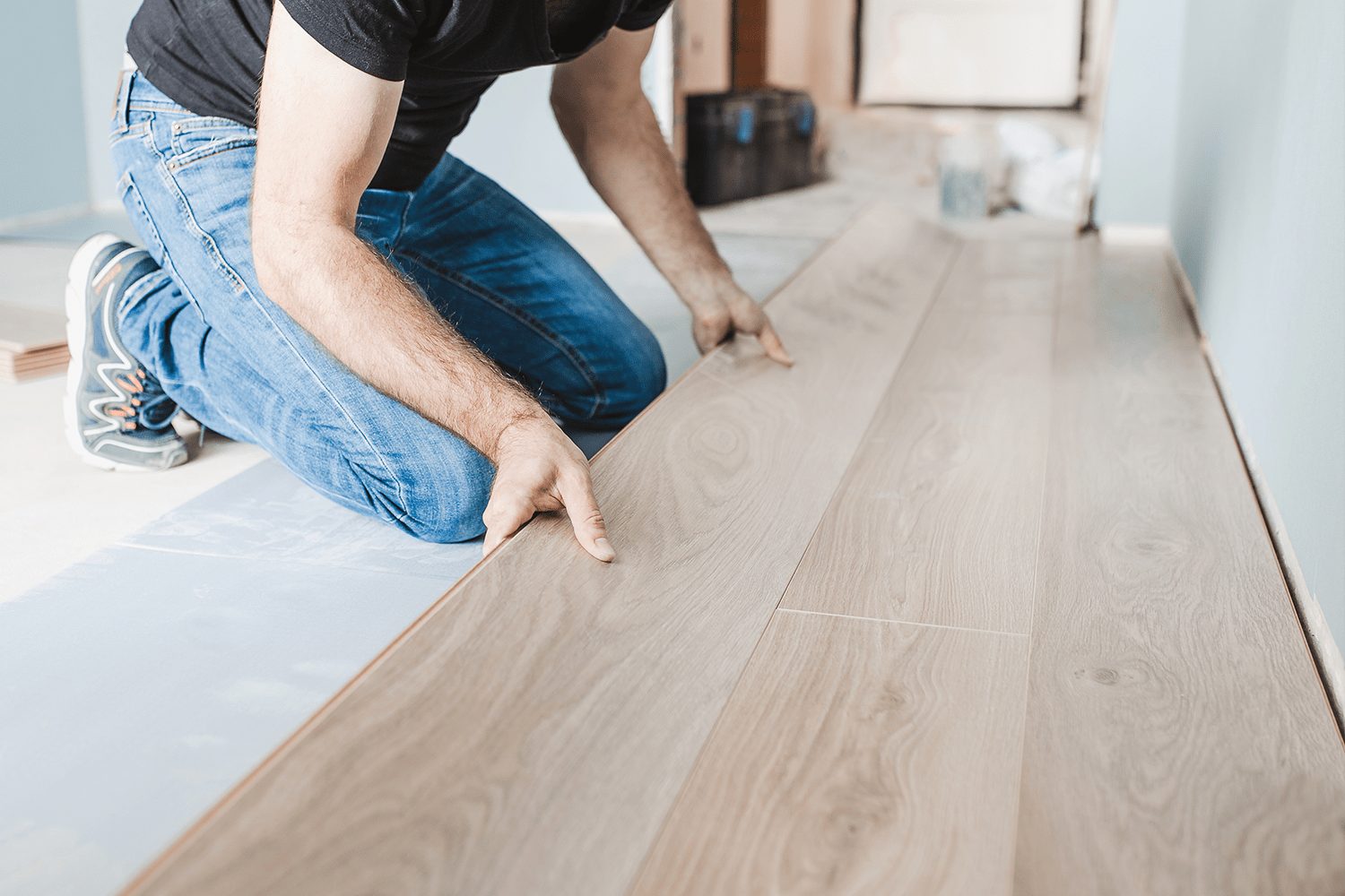 A person kneels, placing wooden floor planks on a subfloor in a room with light blue walls and visible tools in the background.