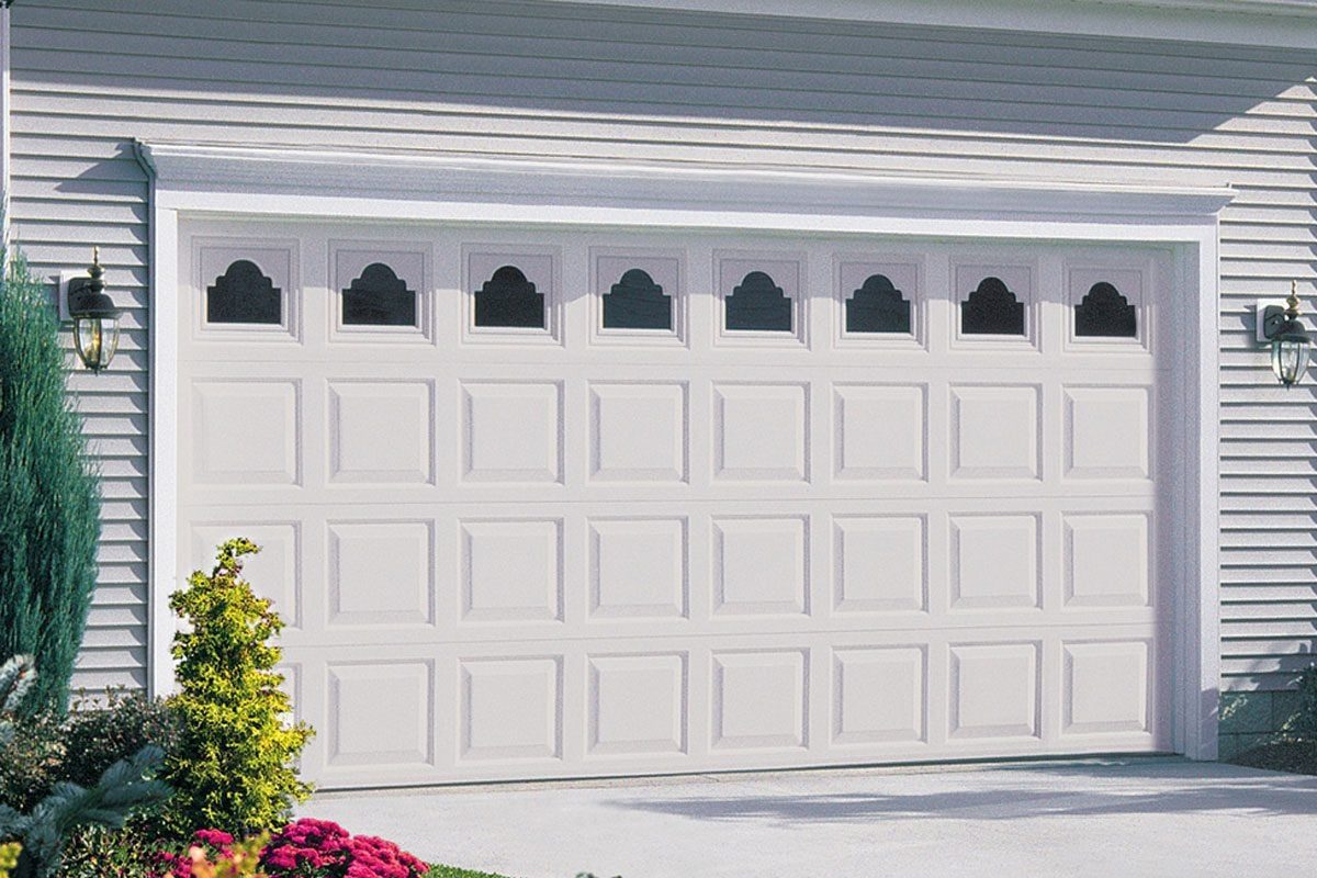 A large, white garage door stands closed, featuring decorative windows on top, amidst a neatly landscaped setting with bushes and flowers in