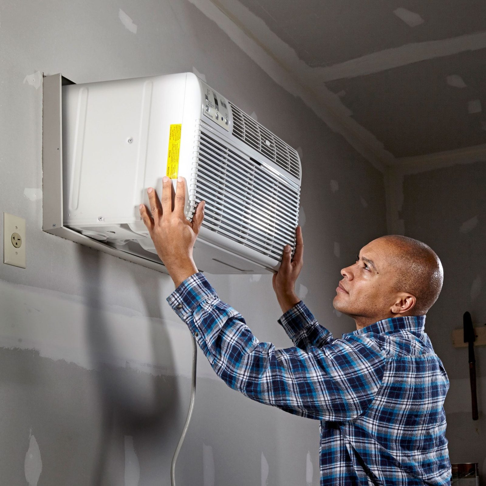 A man installs a window air conditioner unit in a partially finished room with drywall, focusing intently while adjusting it into place.
