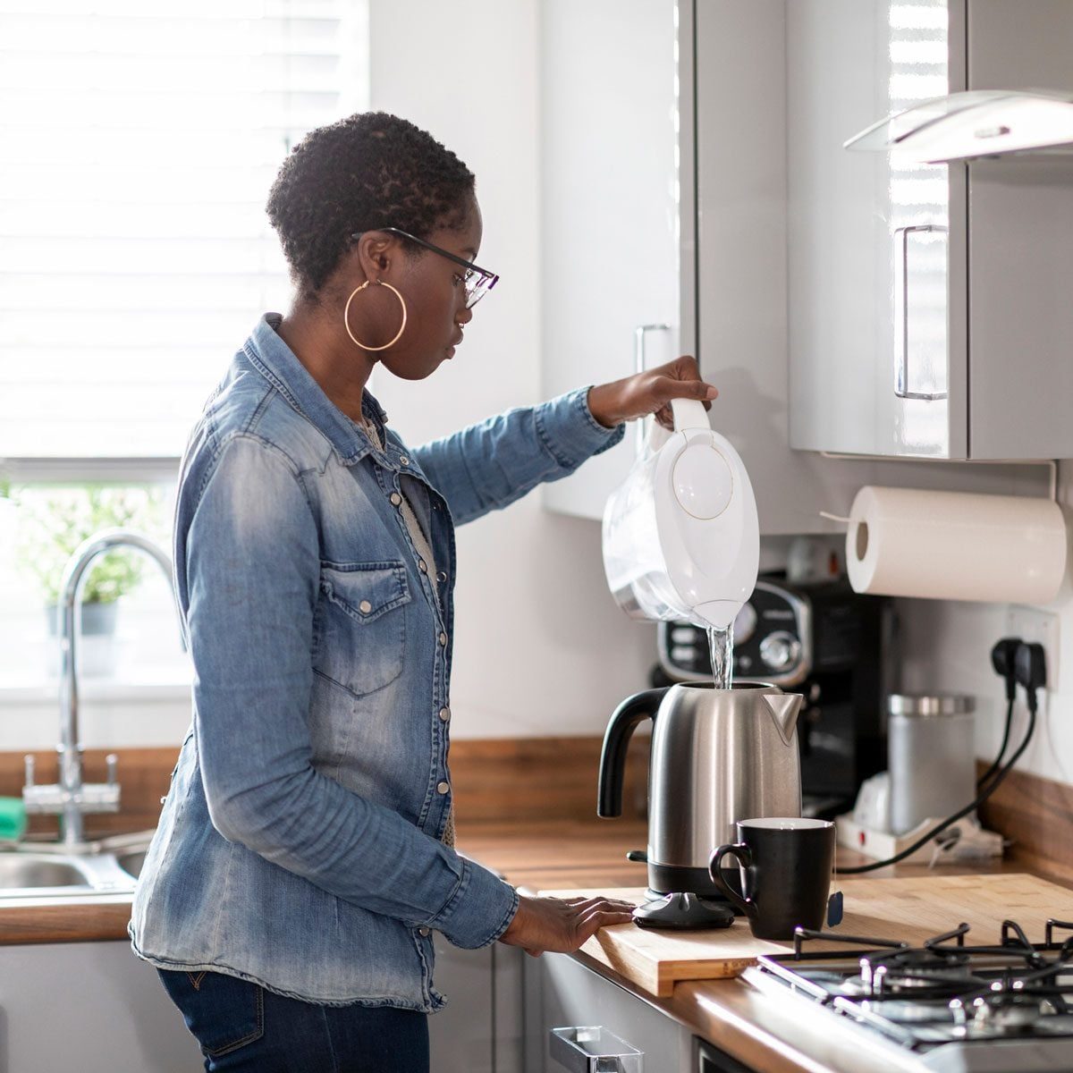 A woman pours water from a kettle into a stainless steel electric kettle on a kitchen countertop, surrounded by a sink and coffee maker.