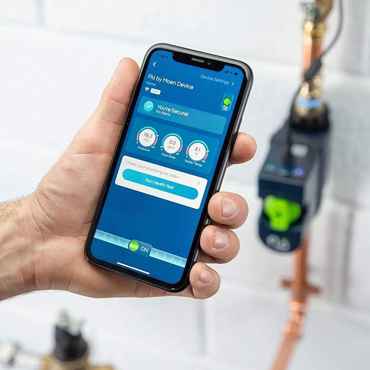 A hand holds a smartphone displaying a plumbing monitoring app, showing security status and water metrics, in a brightly lit room with pipes