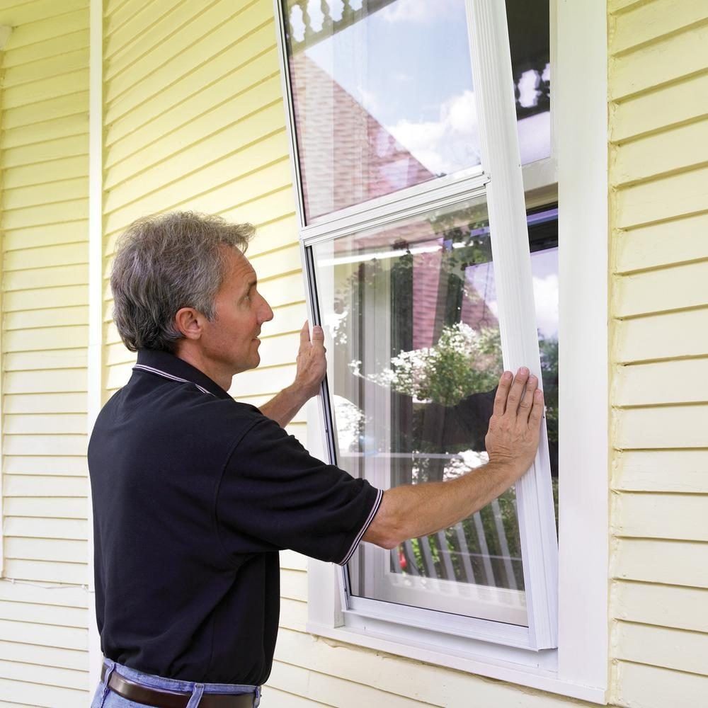 A man is installing a window screen on a yellow house, using both hands to position it against the open window, surrounded by greenery.