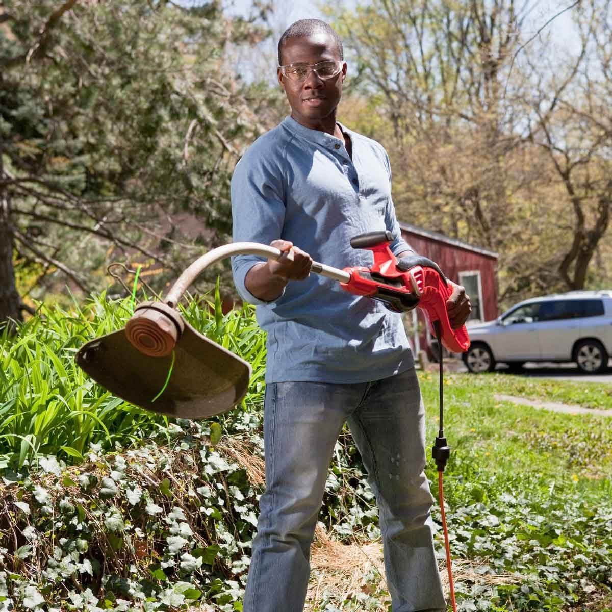 A man holds a lawn trimmer, preparing to cut grass in a garden. Lush green plants surround him, with a red shed and a vehicle in the backgro