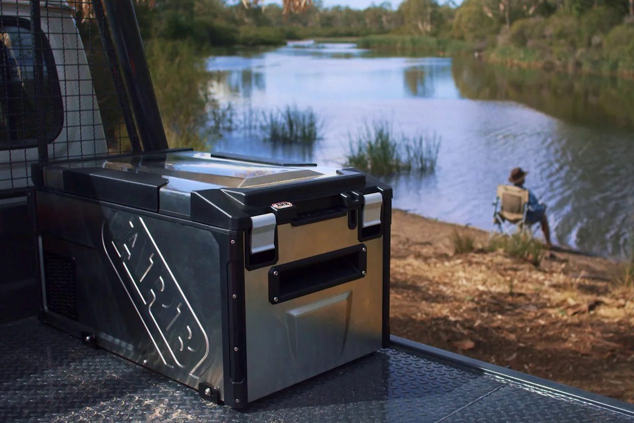A cooler rests on a truck bed, with a tranquil river and a person fishing in the background, surrounded by greenery and nature.