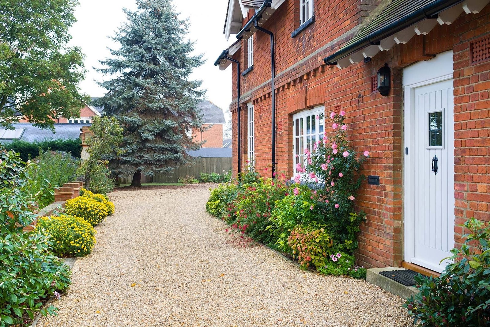 A brick house with a white door is surrounded by flowers and shrubbery along a gravel path, with a large tree in the background.