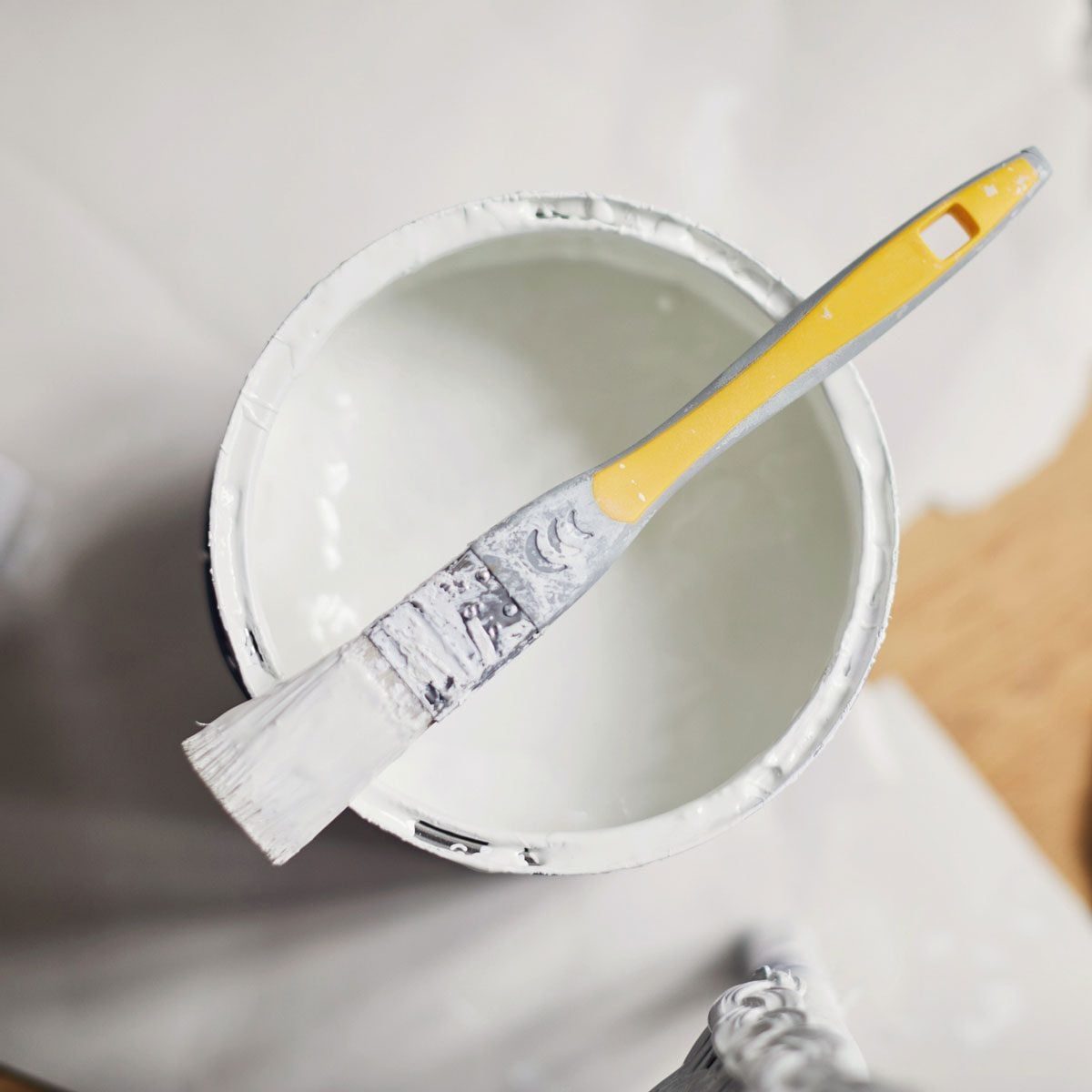 A paintbrush rests on the edge of a white paint can, surrounded by painter's materials on a wooden surface.