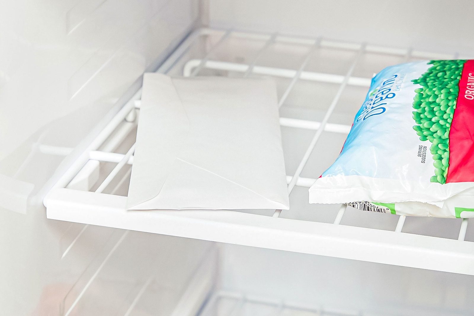 A plain envelope rests on a white refrigerator shelf beside a package of frozen peas, illuminated by the appliance's interior light.