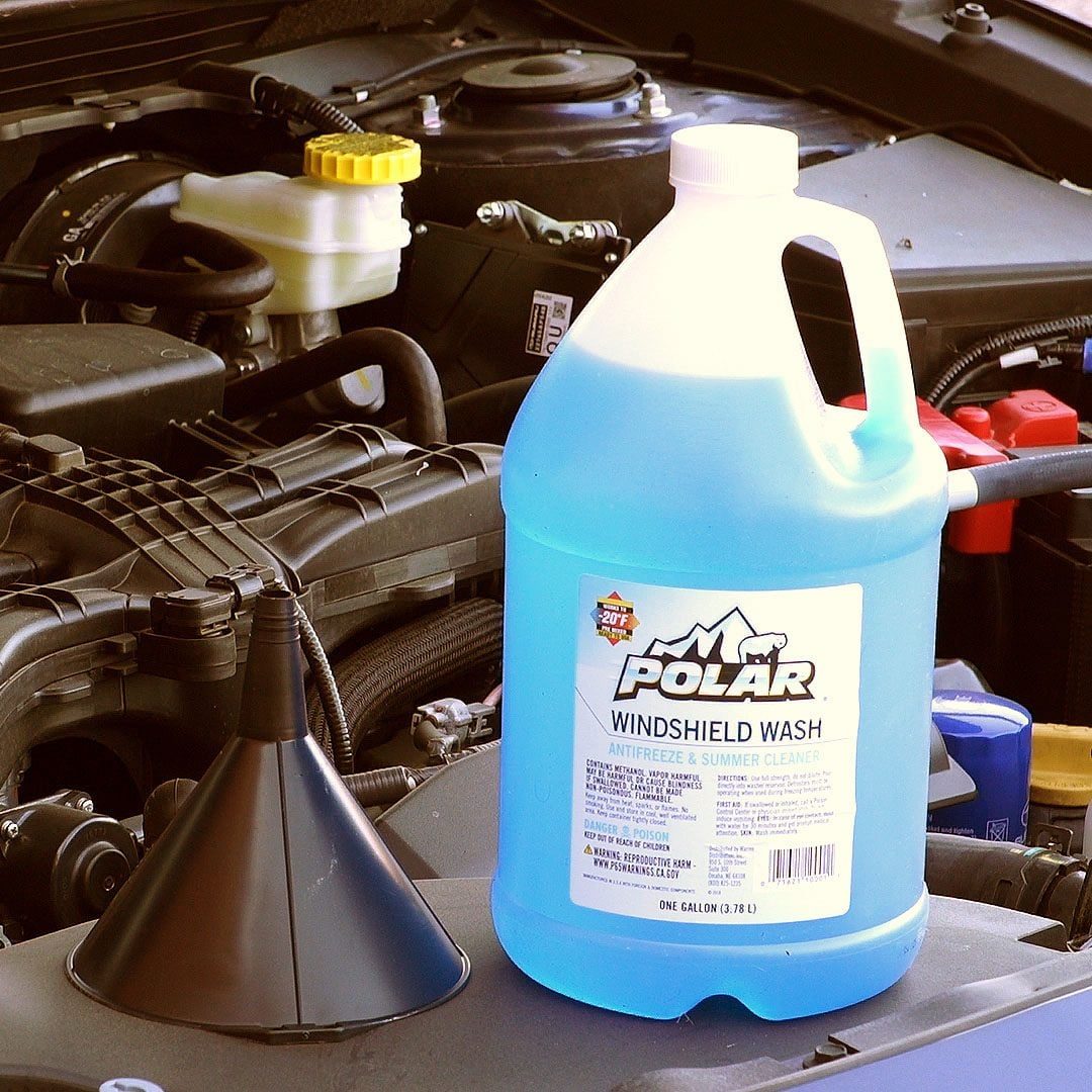 A blue container labeled "POLAR WINDSHIELD WASH" sits beside a funnel, near the open engine compartment of a vehicle.