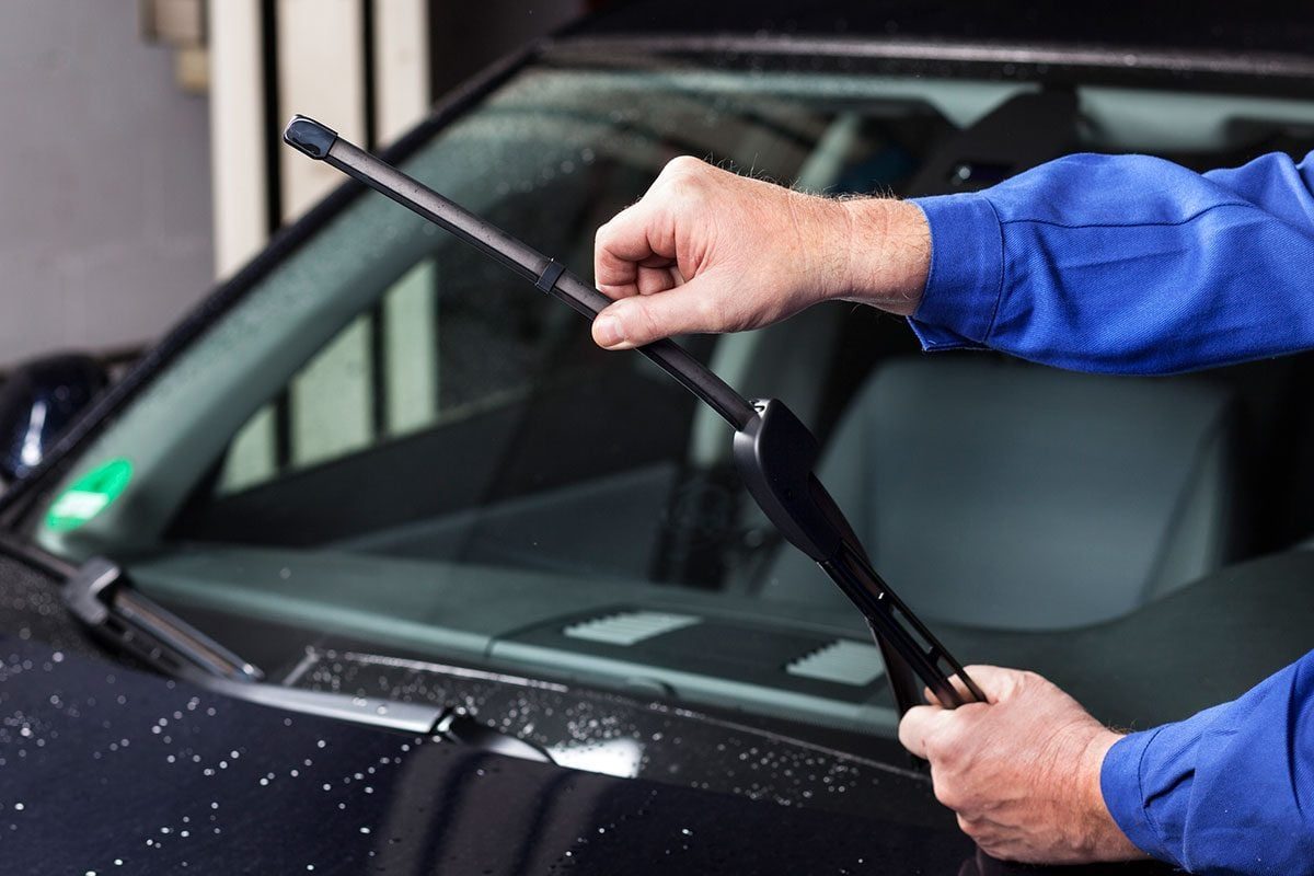 A person is replacing a windshield wiper blade on a car, surrounded by a rainy ambiance with droplets on the vehicle's surface.