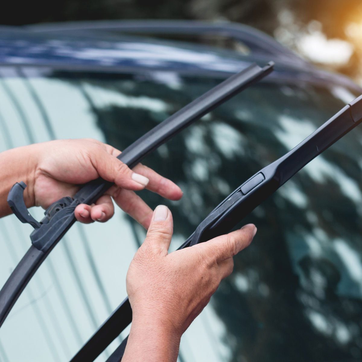 Hands replace windshield wipers on a car. The setting is outdoors, with trees reflecting on the car's glass and a hint of sunlight.