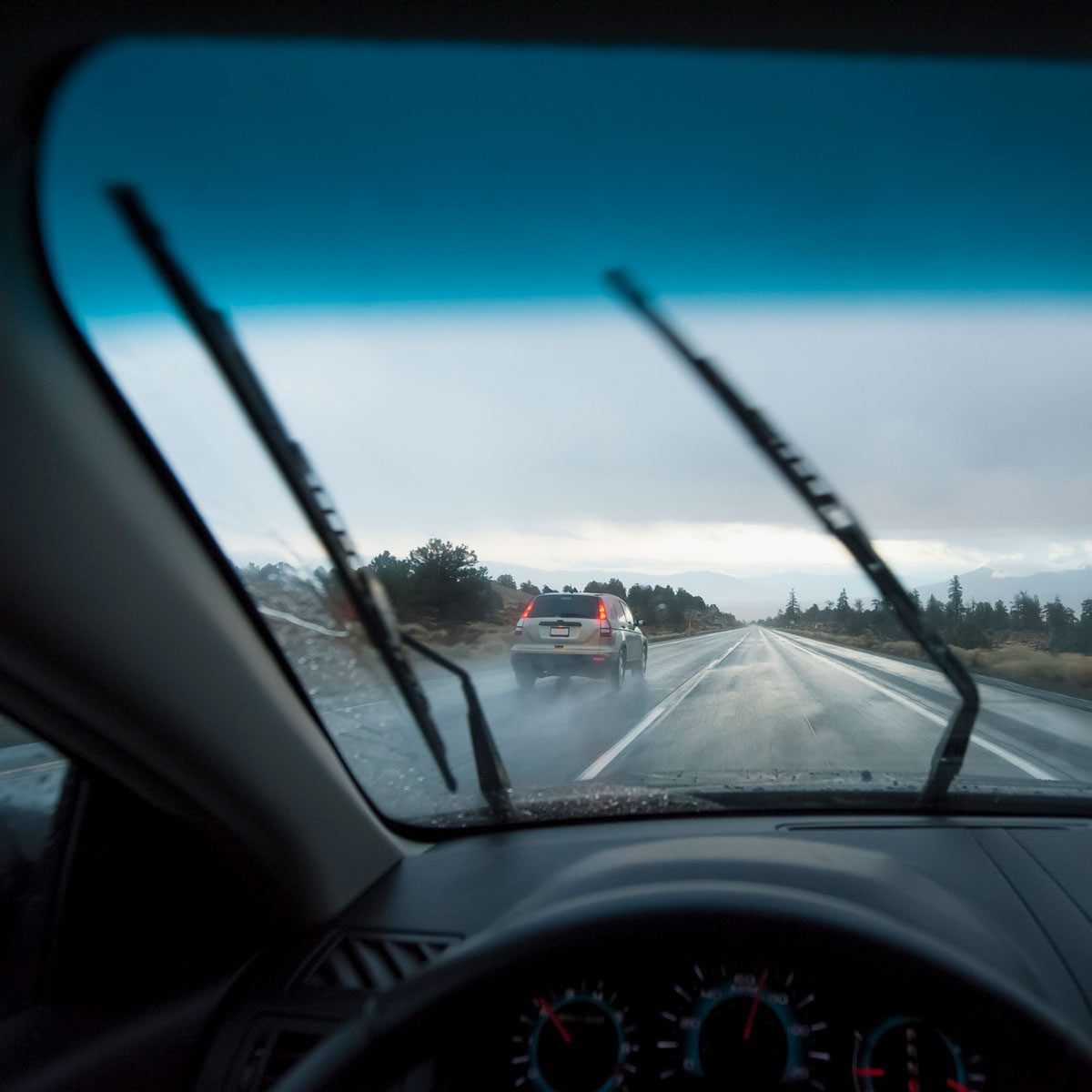 A car is driving on a wet road, while windshield wipers clear rain from the driver’s view. Dark clouds hang overhead, suggesting inclement w