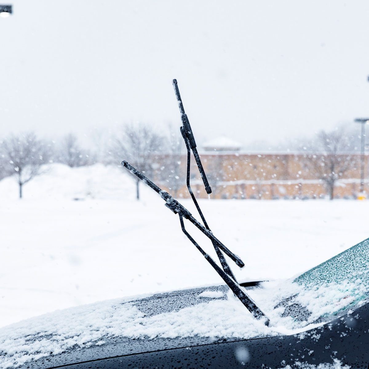 A car with raised windshield wipers is parked in a snowy lot, surrounded by falling snow and mounds of accumulated snow in the background.