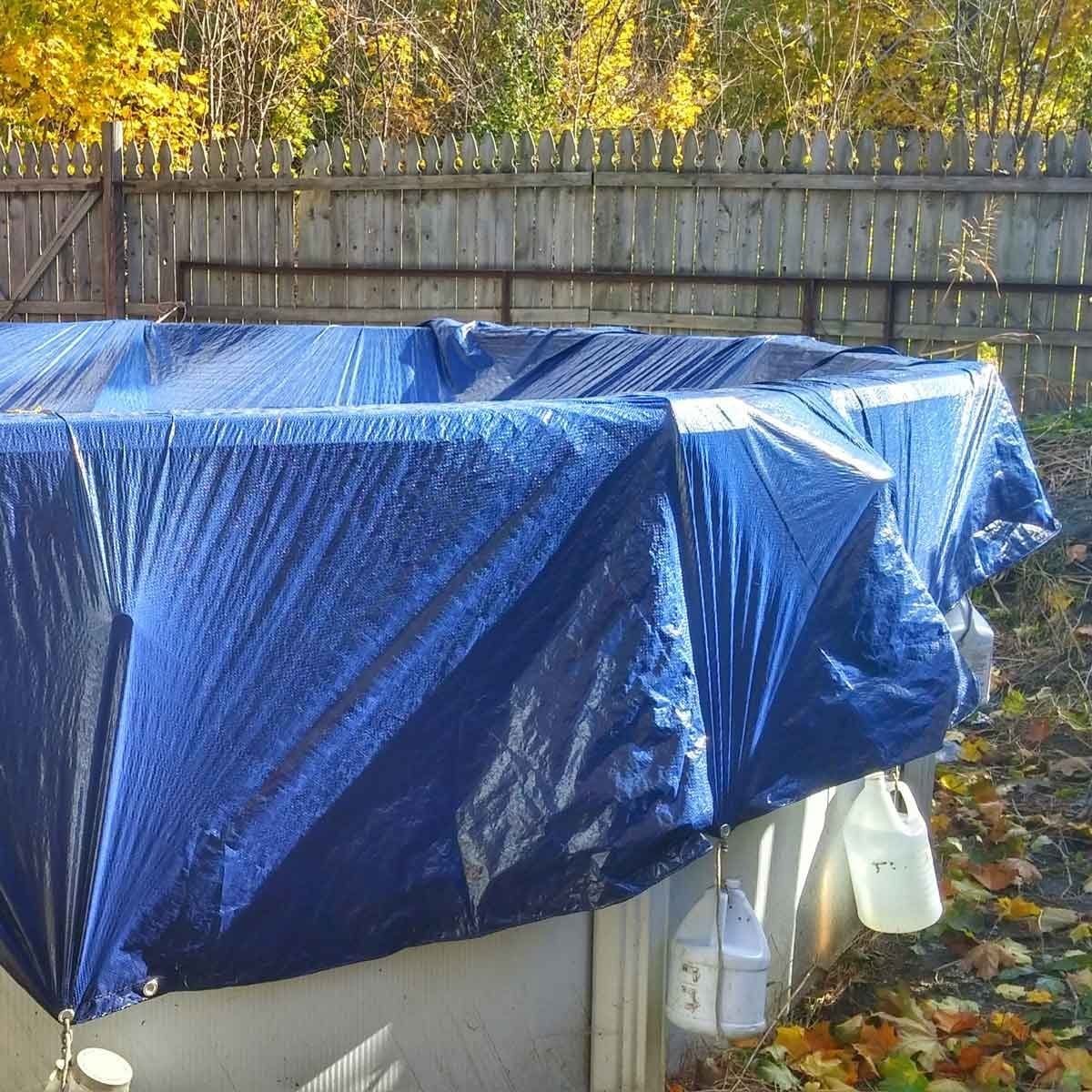 A blue tarp covers several structures in a backyard, securing their contents while surrounded by a wooden fence and autumn-colored foliage.