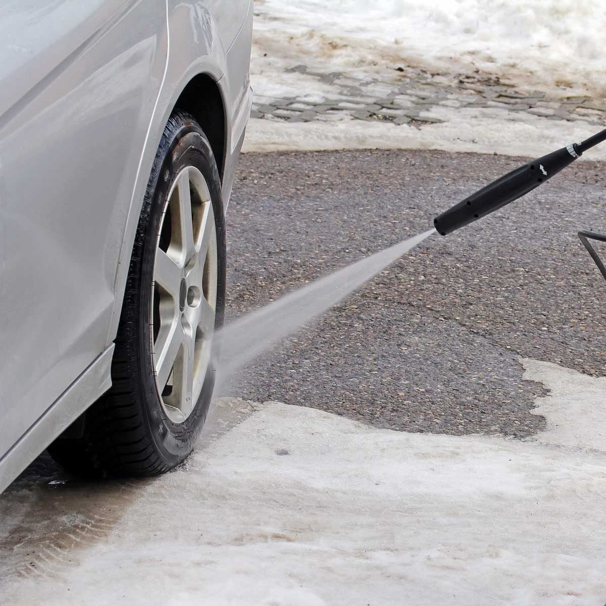A pressure washer sprays water on a car tire, removing dirt, positioned on a wet, icy surface amidst patches of snow and asphalt.