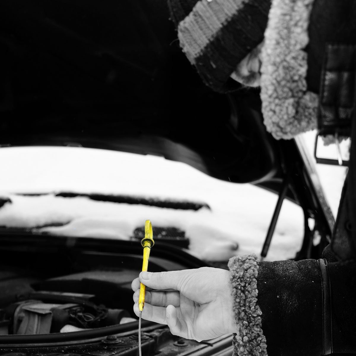 A hand holds a yellow dipstick above an open car hood in a snowy environment, indicating a check on the vehicle's oil level.