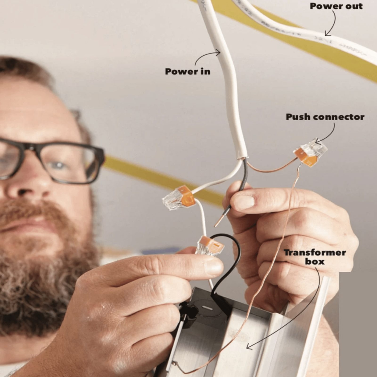A person is connecting electrical wires to a transformer box, surrounded by a ceiling and visible wiring with labels indicating power direct