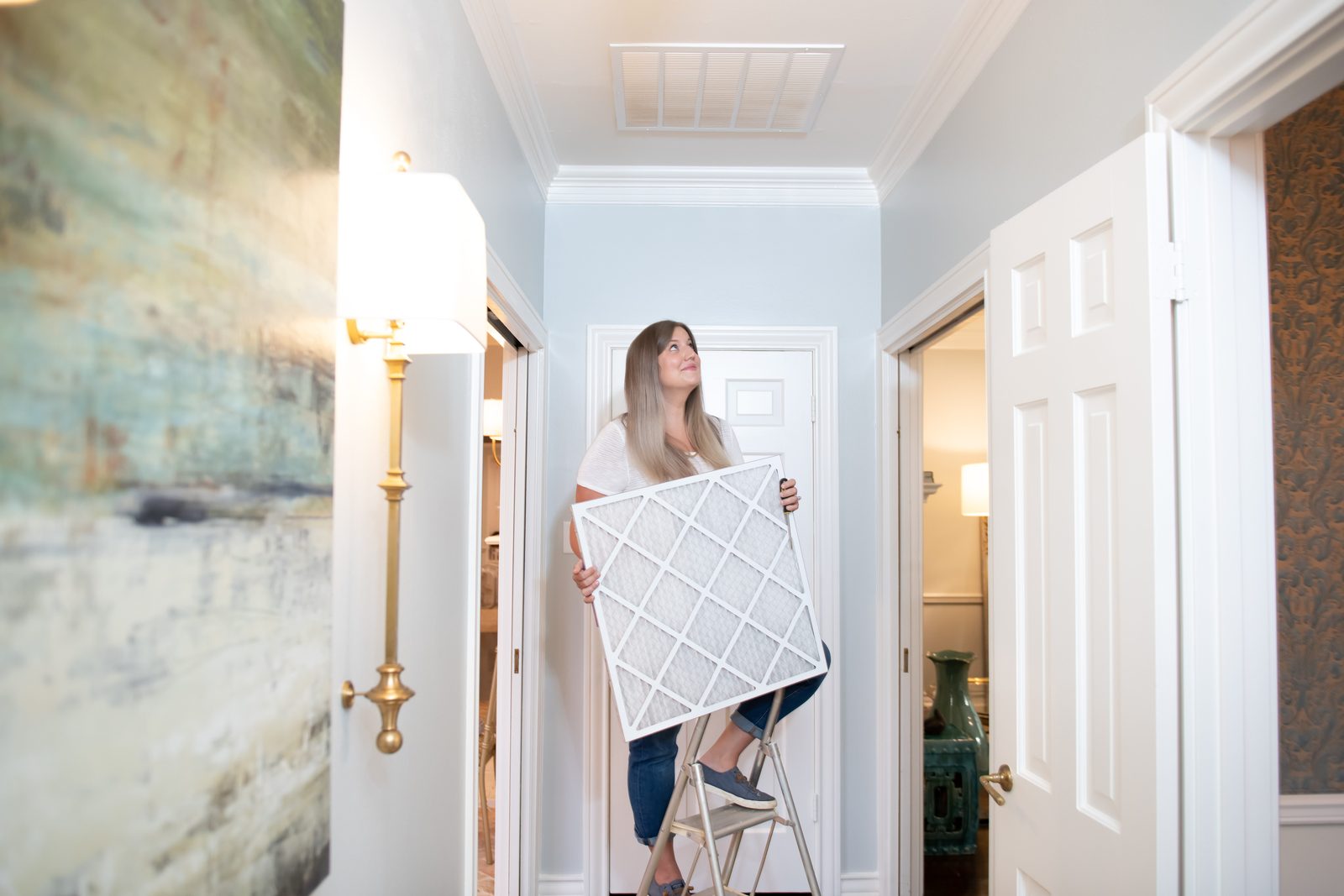 Woman climbing up a small ladder in the corridor holding a duct cover for her home's ventilation system