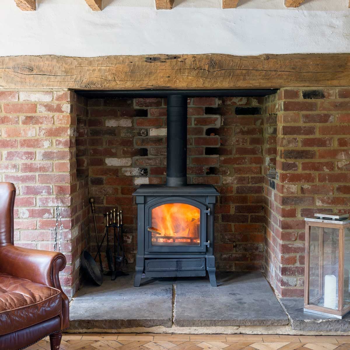 A black stove emits flames in a brick fireplace, surrounded by a wooden beam, a brown leather chair, and a glass lantern on a stone floor.
