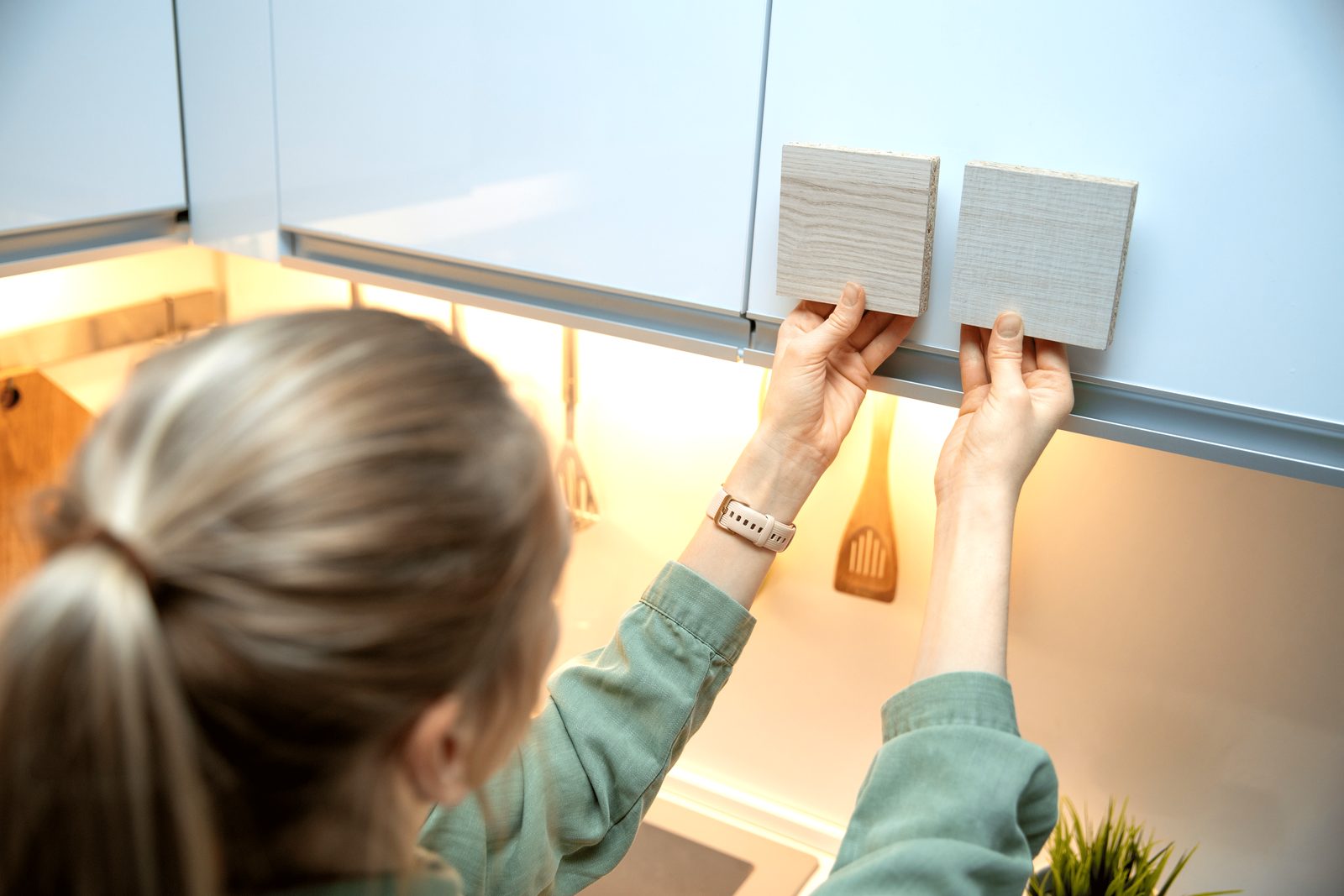 A person holds two wooden samples up to a cabinet in a kitchen, assessing their color and texture under warm lighting.
