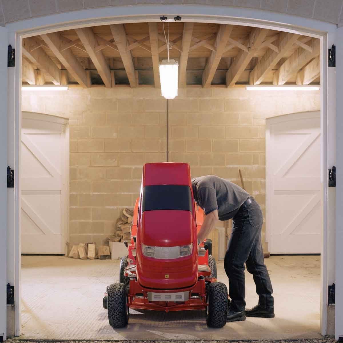 A person leans over a red lawn mower, inspecting it inside a well-lit garage with exposed beams and wooden logs stacked nearby.