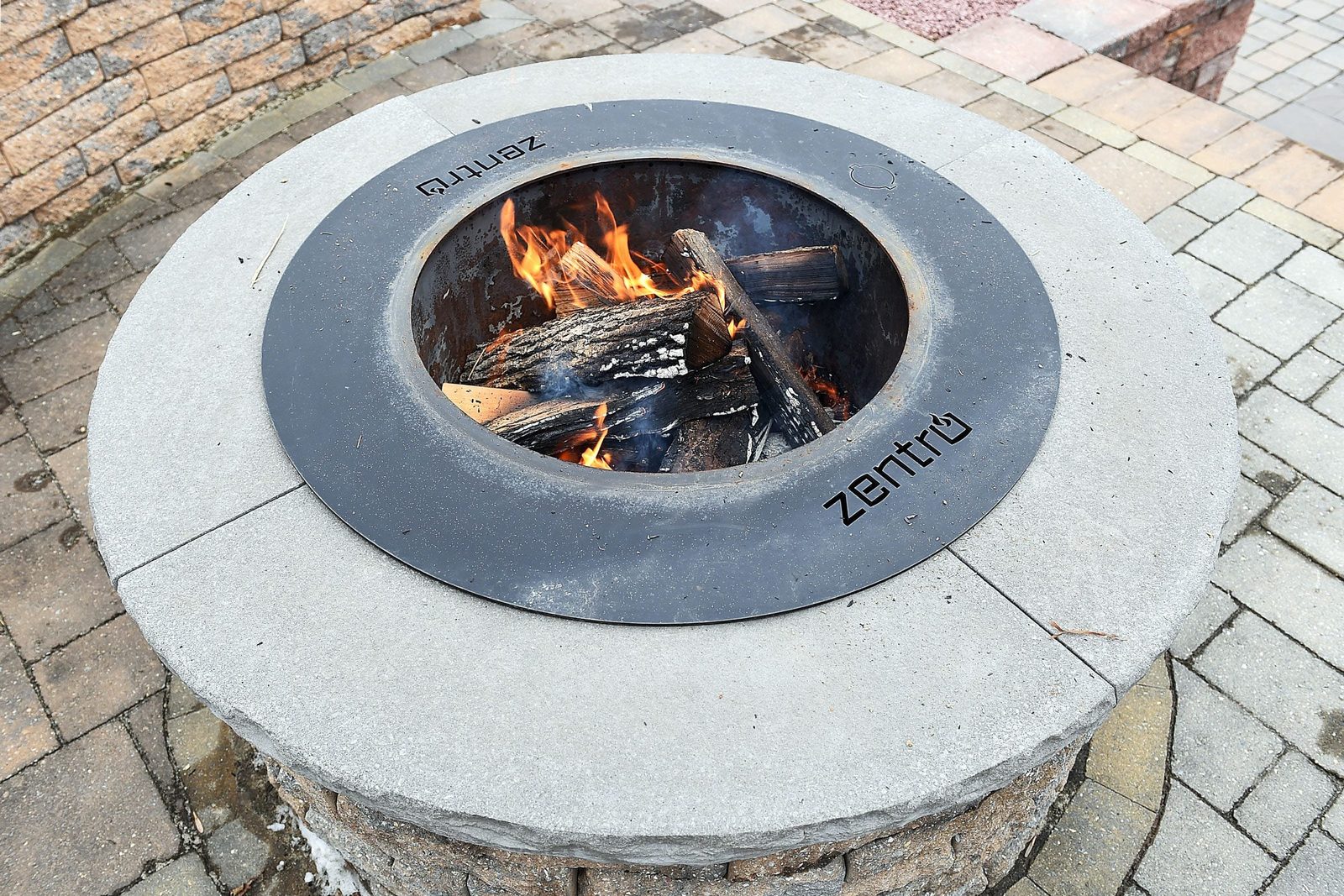 A circular fire pit contains burning logs, emitting flames, placed on a stone patio surrounded by paved walkways.