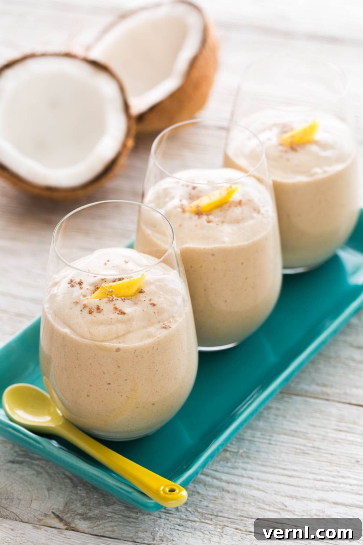 Κρεμώδες smoothie καρύδας-μάνγκο σε 3 ποτήρια πάνω σε ένα μπλε πιάτο.
