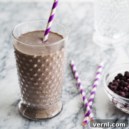 Ένα smoothie σοκολάτα-μύρτιλο σε ένα ποτήρι με μωβ ριγέ καλαμάκια και μύρτιλα δίπλα.