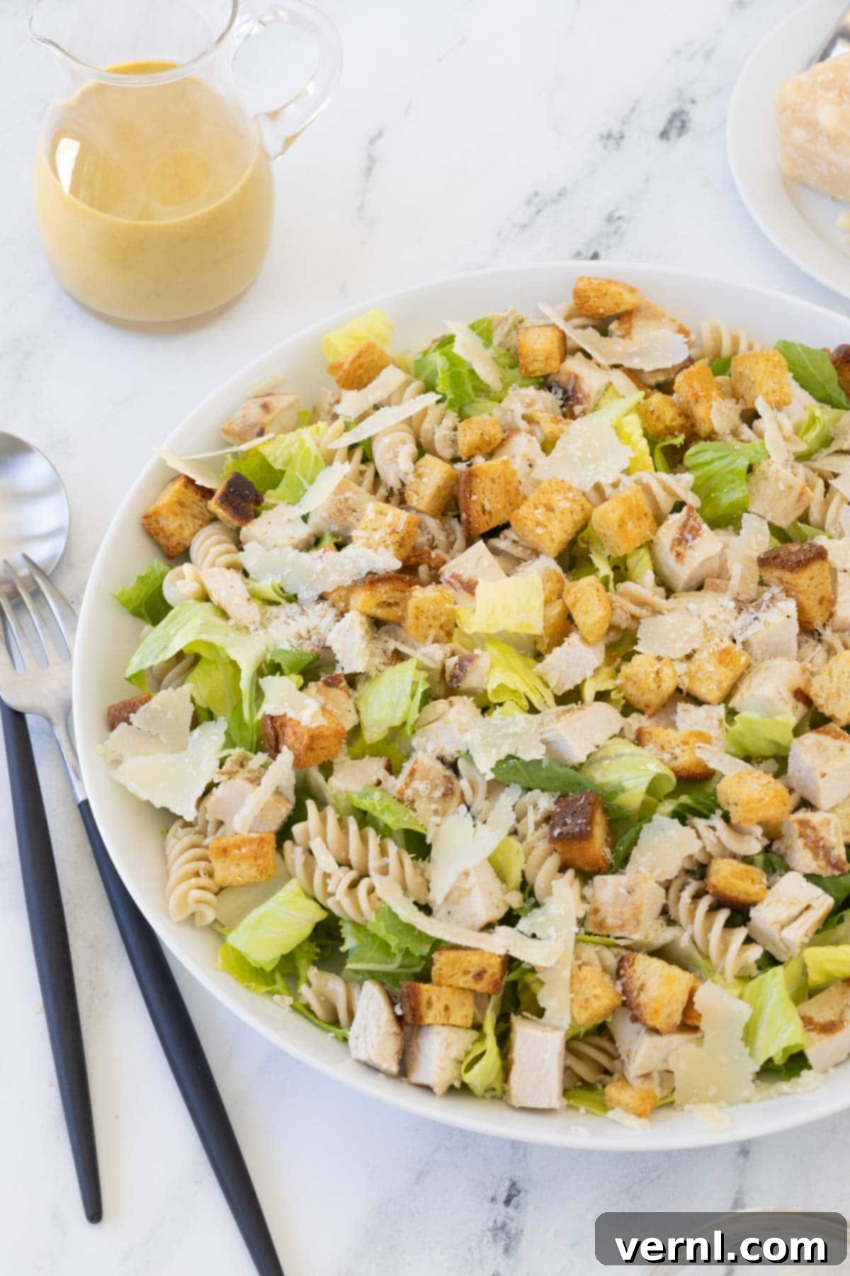 Σαλάτα Ζυμαρικών Καίσαρα με Κοτόπουλο 2 A large shallow bowl of caesar pasta salad with romaine, curly pasta, croutons, chicken and dressing in a small pitcher.