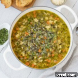Μια λευκή κατσαρόλα με σούπα γαλοπούλας και άγριου ρυζιού σε γκρι ξύλινη σανίδα με μυρωδικά και τυρί παρμεζάνα στο πλάι.