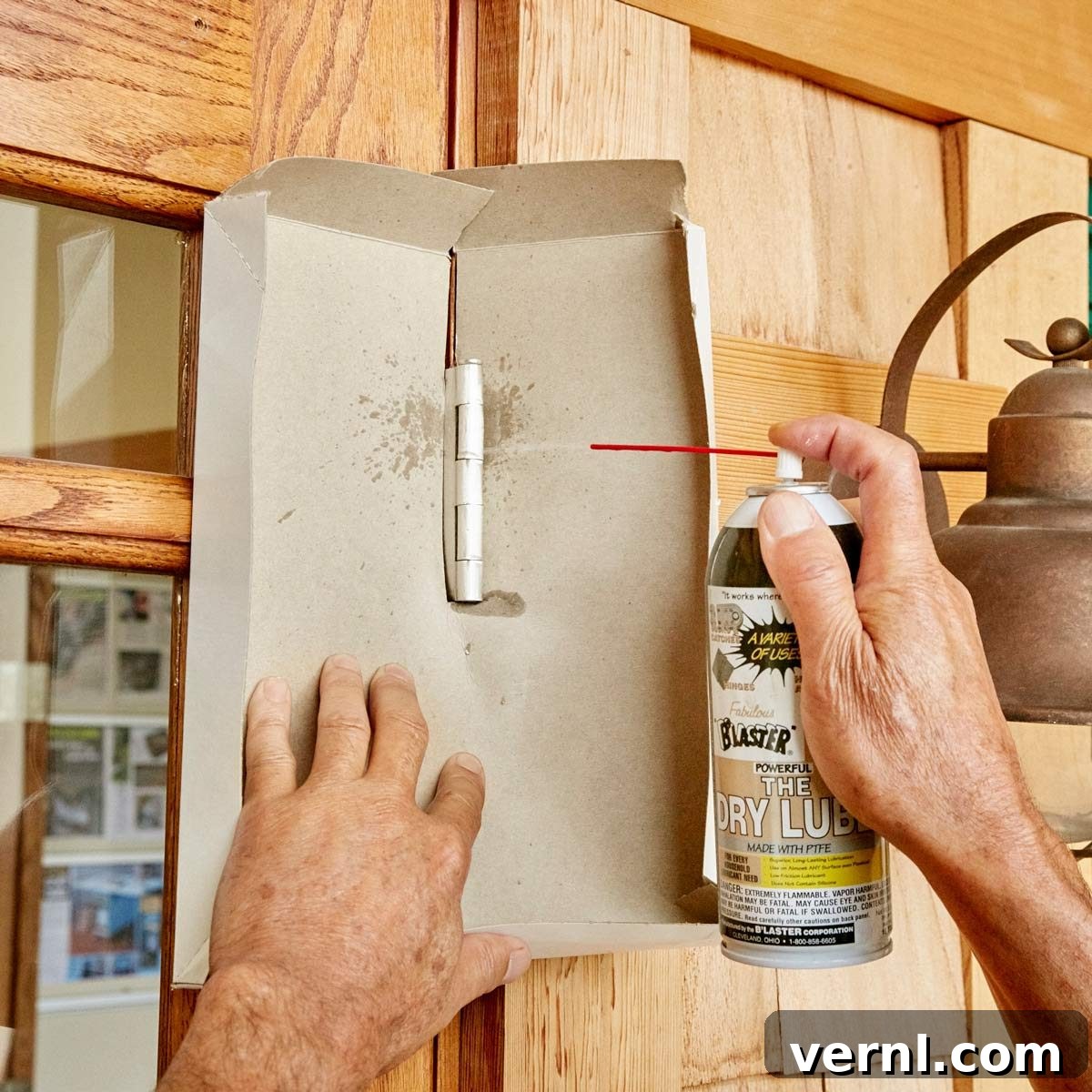 Person spraying dry lubricant on a squeaky door hinge with cardboard protection