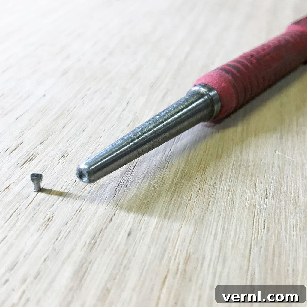 A versatile metal nail set, featuring a pointed tip, lies near a small screw on a beautifully finished wooden surface, symbolizing the final touches of a precise woodworking project.