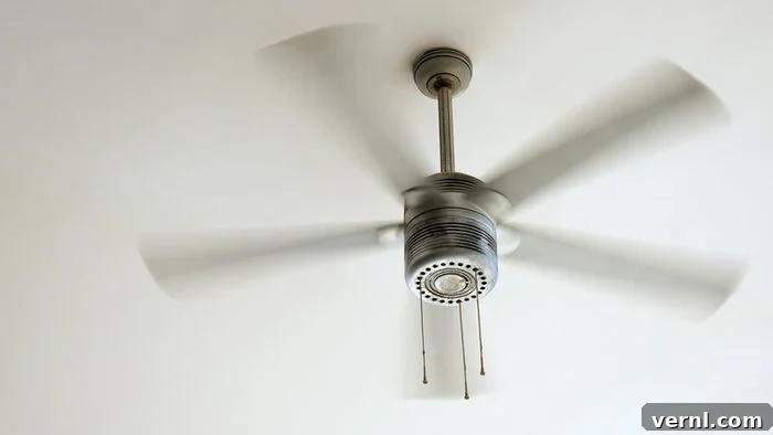 Ceiling Fans: Energy Hogs or Efficiency Heroes 2 A modern ceiling fan with four blades rotating gracefully, creating a gentle breeze in a well-lit living space. The fan is mounted on a white ceiling, blending seamlessly with the room's contemporary decor. Soft, natural light streams in from a nearby window, highlighting the fan's sleek design. This image evokes comfort, efficiency, and modern living.