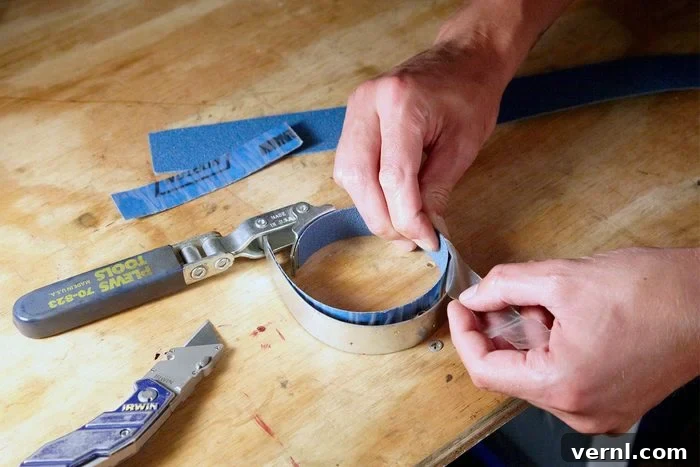 Conquer Stubborn Oil Filters with This Clever Method 3 A close-up image of hands working on a wooden surface with tools. One hand is gripping a piece of blue sandpaper, while the other peels back a layer from it. A utility knife and tape measure are on the table nearby.