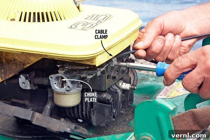 Quick Fix for a Jammed Mower Throttle Cable 3 Close-up of a person's hand using a screwdriver to adjust a cable clamp near the choke plate of a small engine. The engine has a yellow cover, and the green base is visible in the foreground. Labels identify the cable clamp and the choke plate.