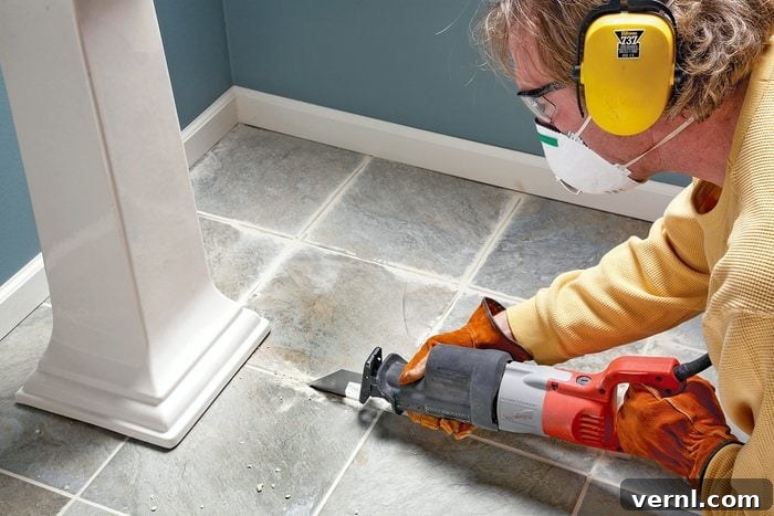 A person wearing safety goggles, ear protection, gloves, and a mask uses an electric saw to cut into a tiled floor near the base of a pedestal table. The person appears focused on the task in a corner of a room.