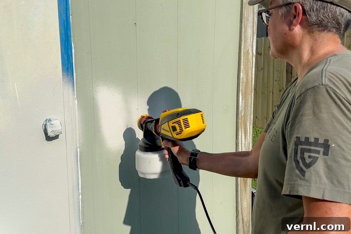 Wagner Flexio 3500: My Secret to a Flawless Paint Job 4 A person wearing a beige cap, gray t-shirt, and glasses uses a yellow and black paint sprayer to paint a green vertical surface. The person is standing outdoors next to a wooden structure.