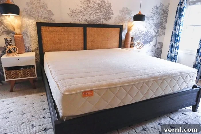 Happsy Mattress Review Organic Comfort for Allergy Sufferers 3 A serene bedroom features a large bed with a light-colored Happsy organic mattress on a dark wood frame. The room boasts tree-patterned wallpaper, two elegant hanging pendant lights, and a minimalist white nightstand adorned with wicker baskets on the left. Soft blue patterned curtains elegantly frame the window on the right, creating a peaceful ambiance.