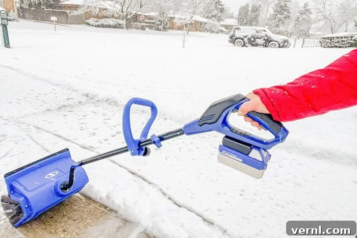 A person in a red jacket uses a blue cordless snow shovel to clear snow from a sidewalk on a snowy day; houses and a car are visible in the snowy background.