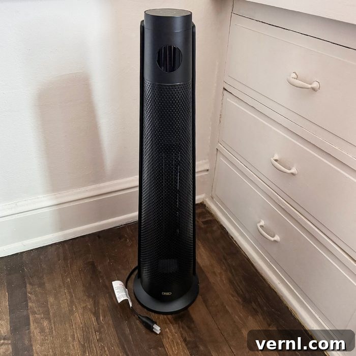 Dreo Uzay Isıtıcısı: Oda Isıtmada Bir Numaralı Tercihimiz 3 A tall, black tower fan or heater stands on a wooden floor next to a white dresser and wall outlet, with its power cord lying on the floor.