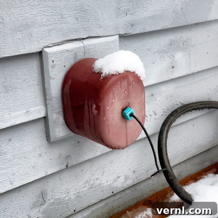 Banish Burst Pipes: Winterize Your Spigot 2 A red outdoor faucet cover with a cable coming out, mounted on a gray house wall. Snow is resting on top of the cover and on the ground below.