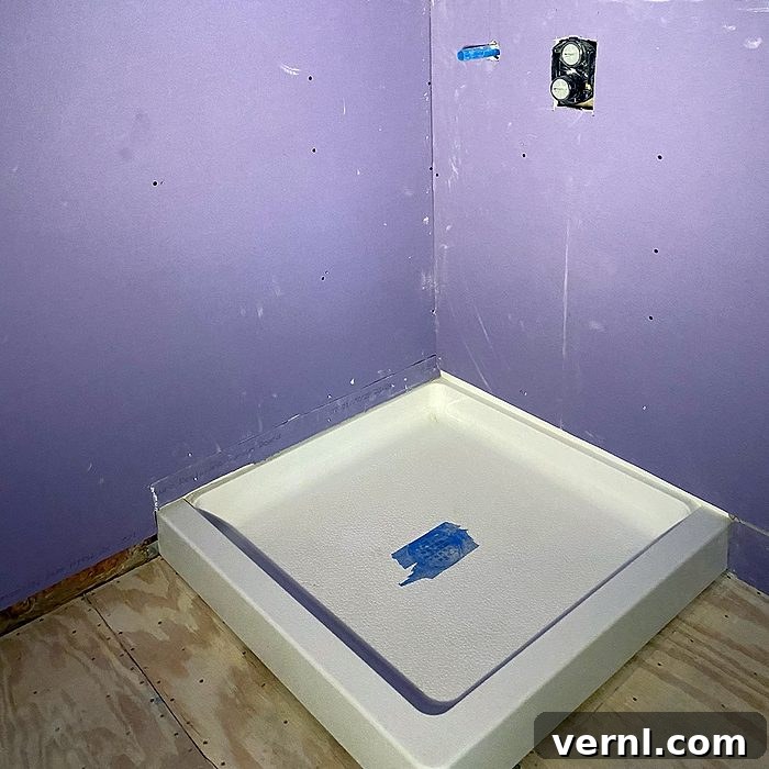 Why I Stopped Renovating for Style and Focused on Function 2 A purple-walled bathroom under construction features an empty white shower pan on a plywood floor. Blue tape covers the drain, and plumbing fixtures are visible.